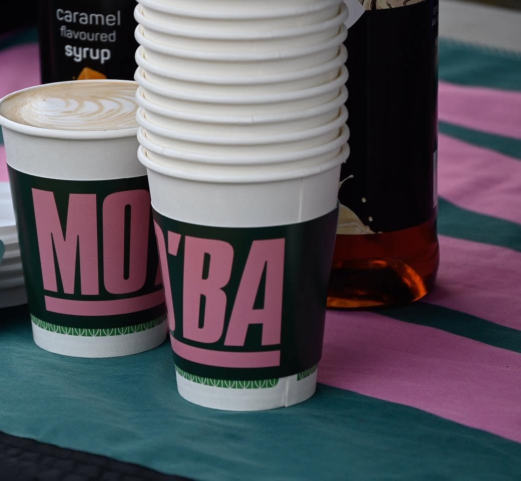 A MO'BA cup filled with a cappuccino and rosetta latte art, standing beside a stack of cups.