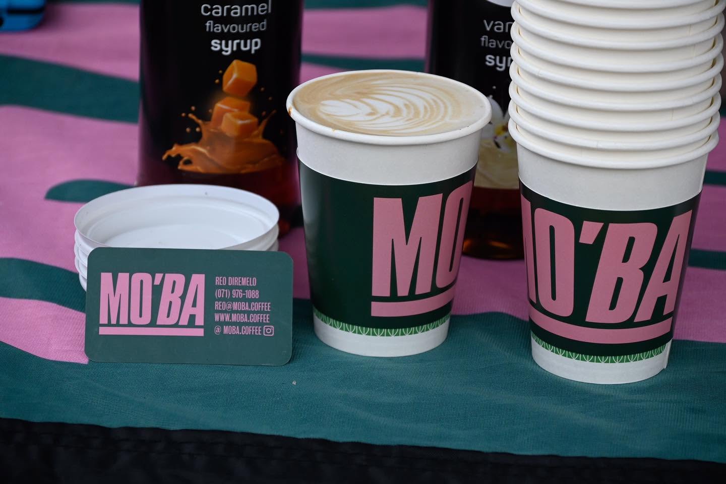 A MO'BA cup of cappuccino with latte art beside a stack of cups and a MO'BA business card.