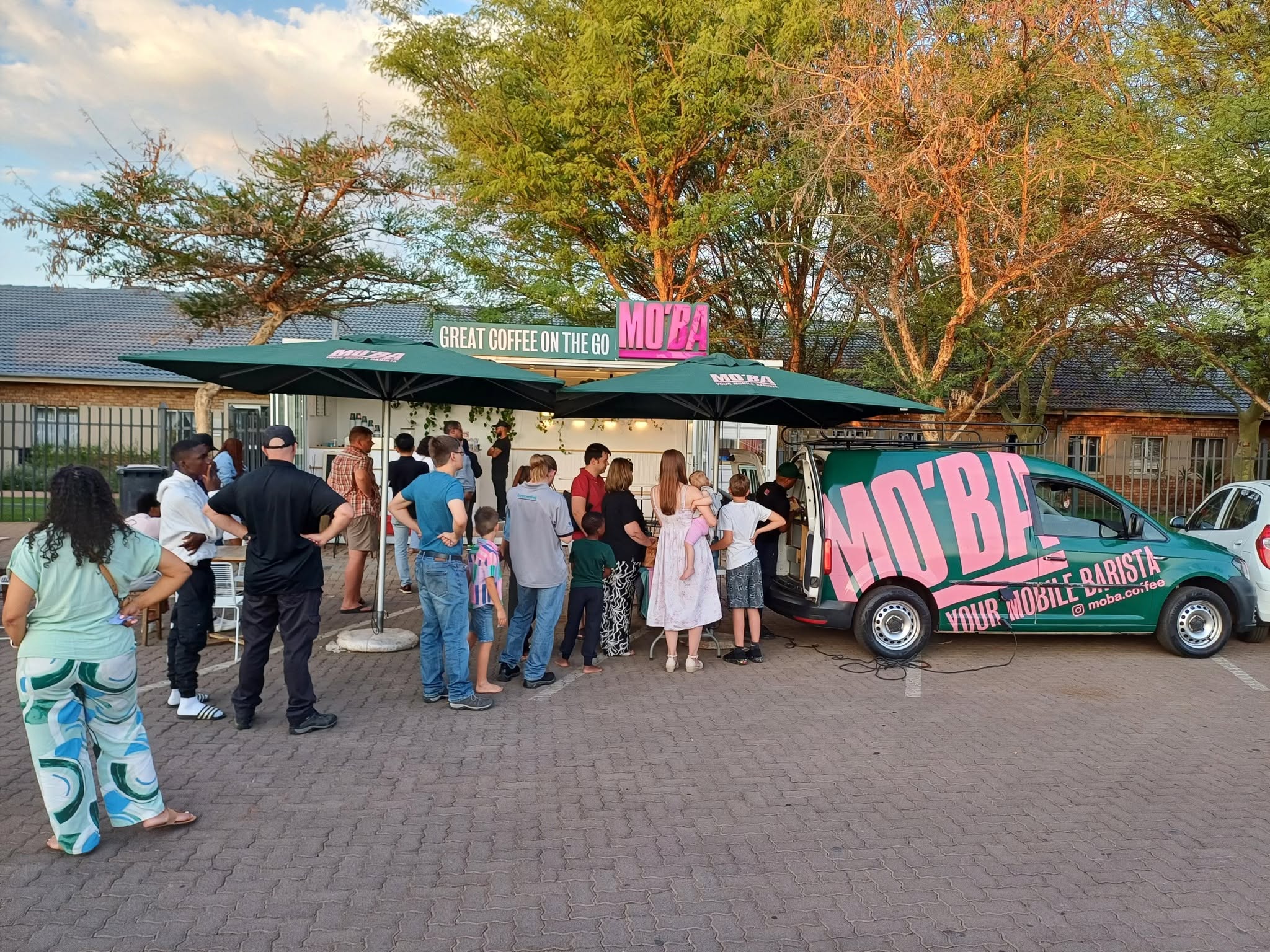 A queue of guests, including families and children, waiting at the MO'BA van beside a container coffee bar at dusk.