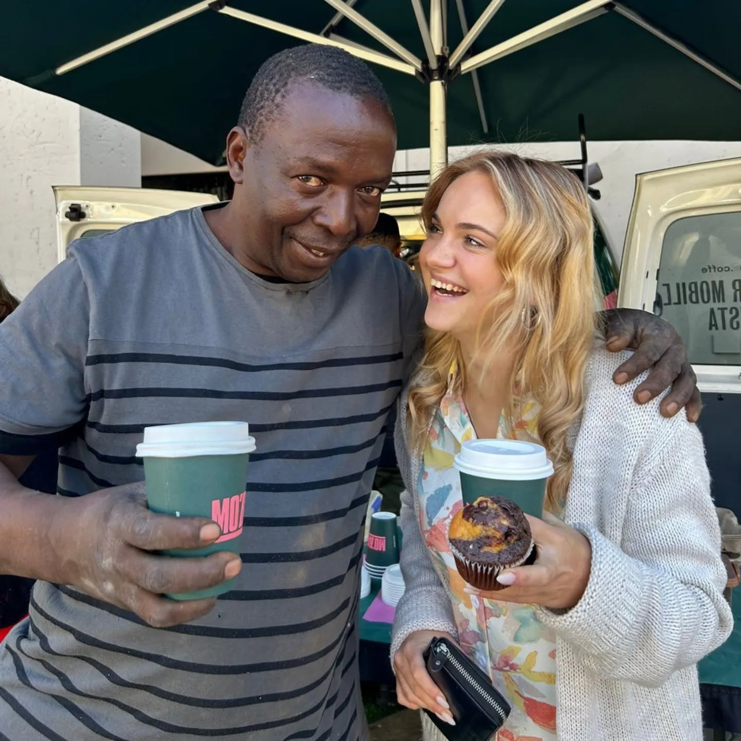 An older man and a young woman laughing together, each holding a MO'BA coffee cup, with the van behind them.