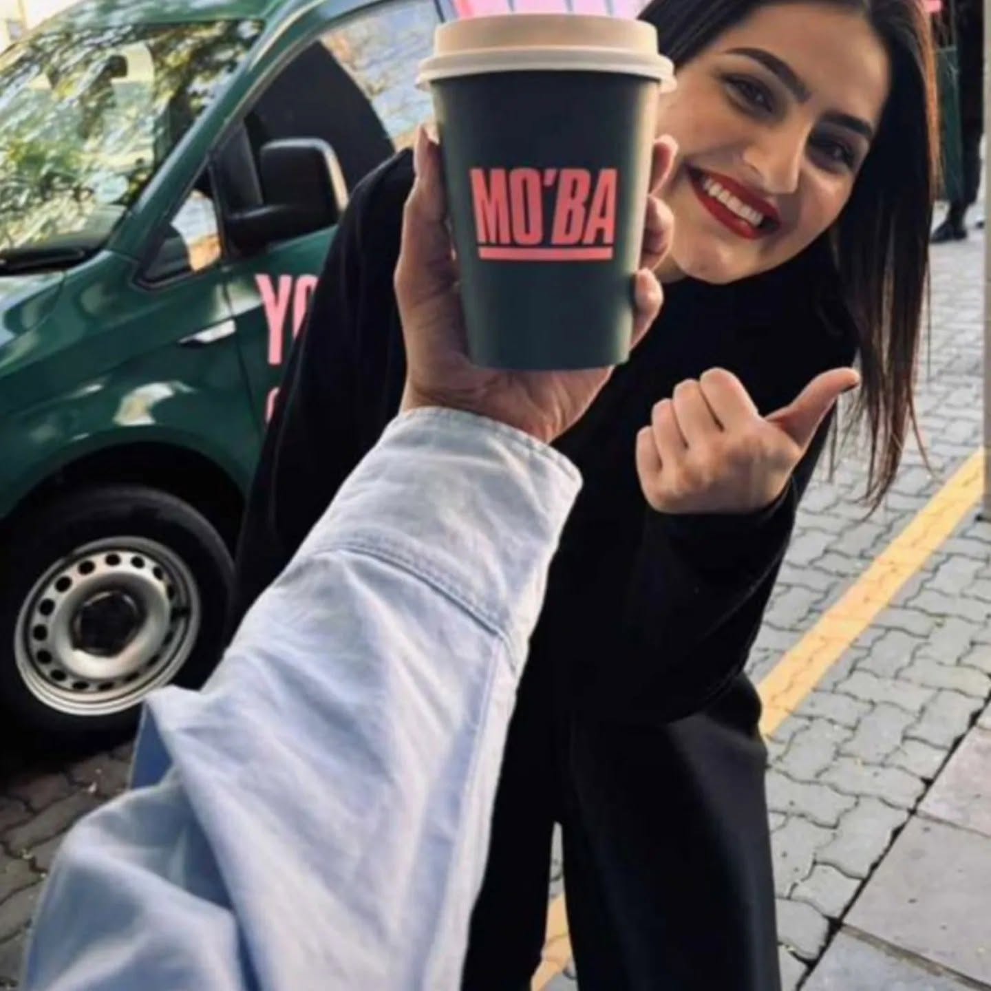 A woman smiling and holding up a MO'BA cup to the camera, with the van in the background.