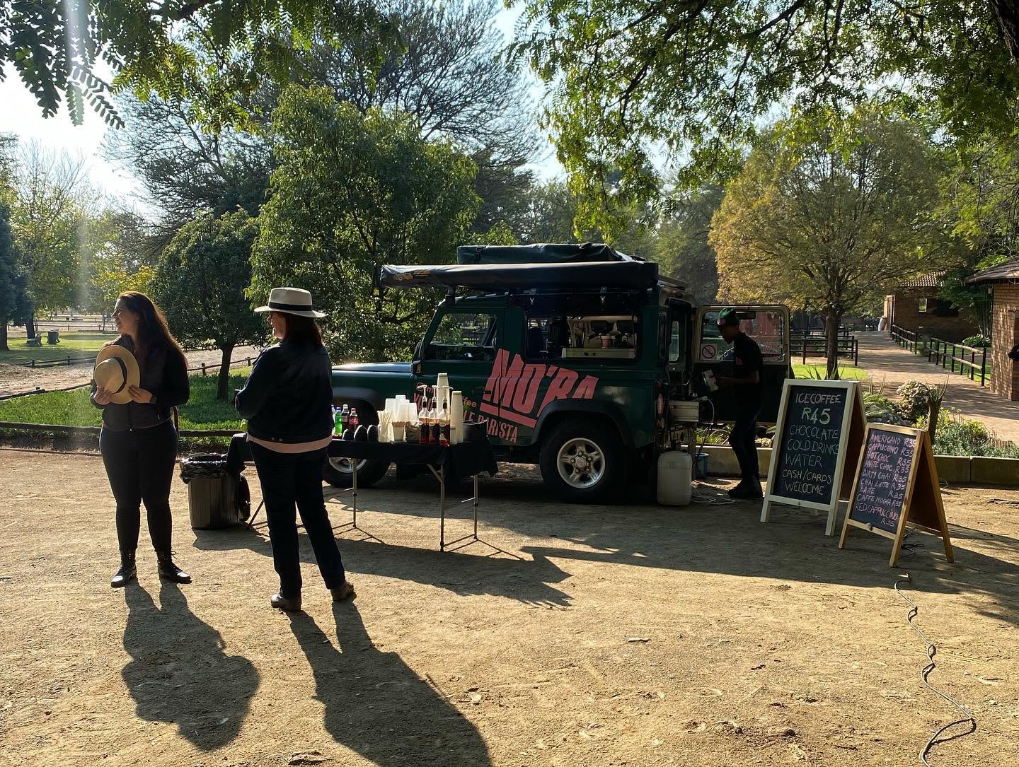 The MO'BA Defender set up under trees in a park, with two guests waiting and a barista serving from the vehicle.