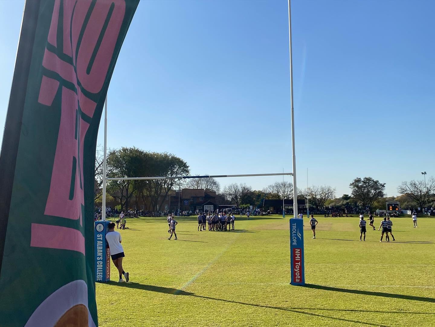 A pink and green MO'BA flag beside a school rugby field mid-match.