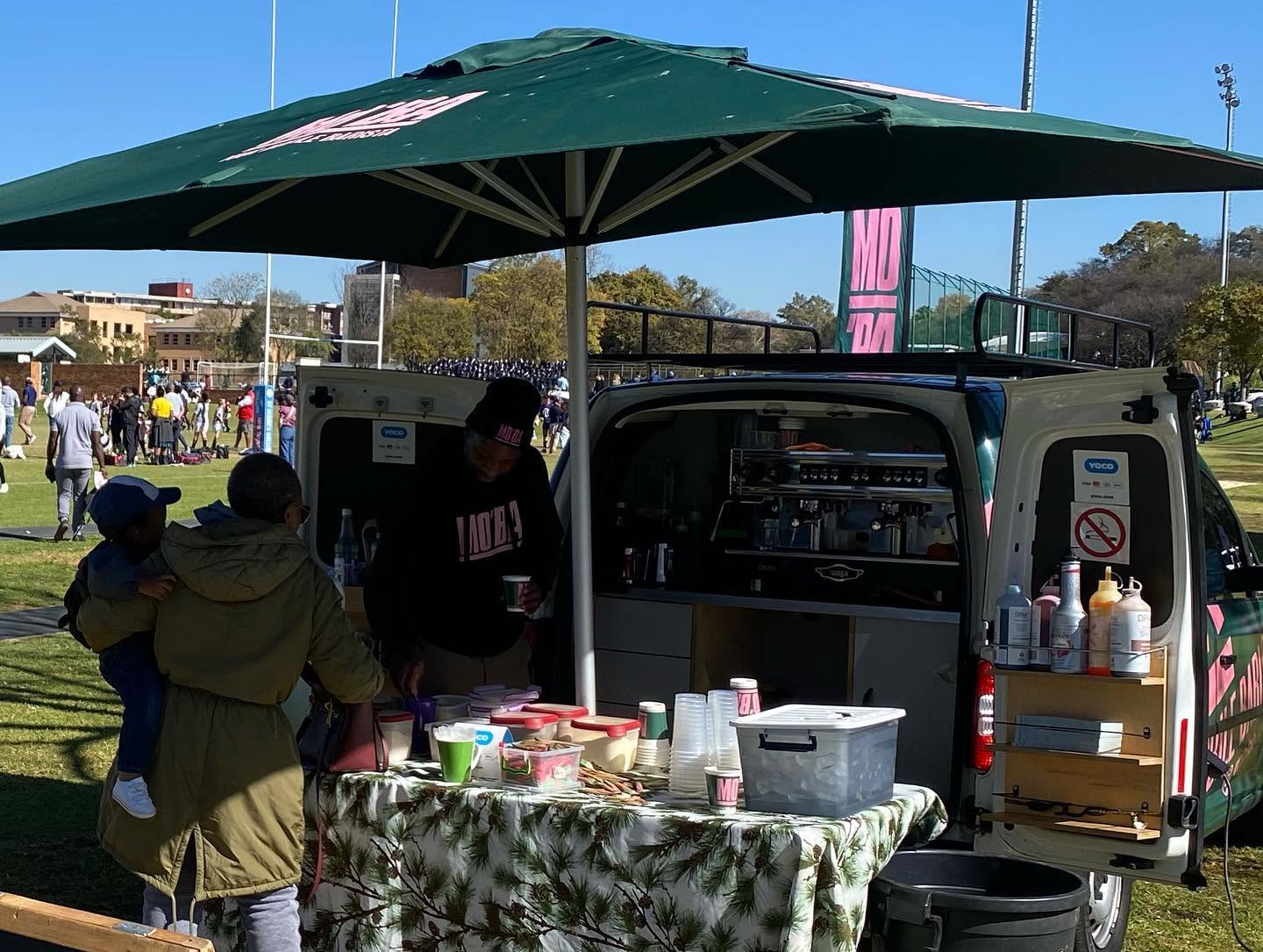 A parent holding a child at the MO'BA Defender's coffee counter, set up beside a school sports field with a rugby match in progress.