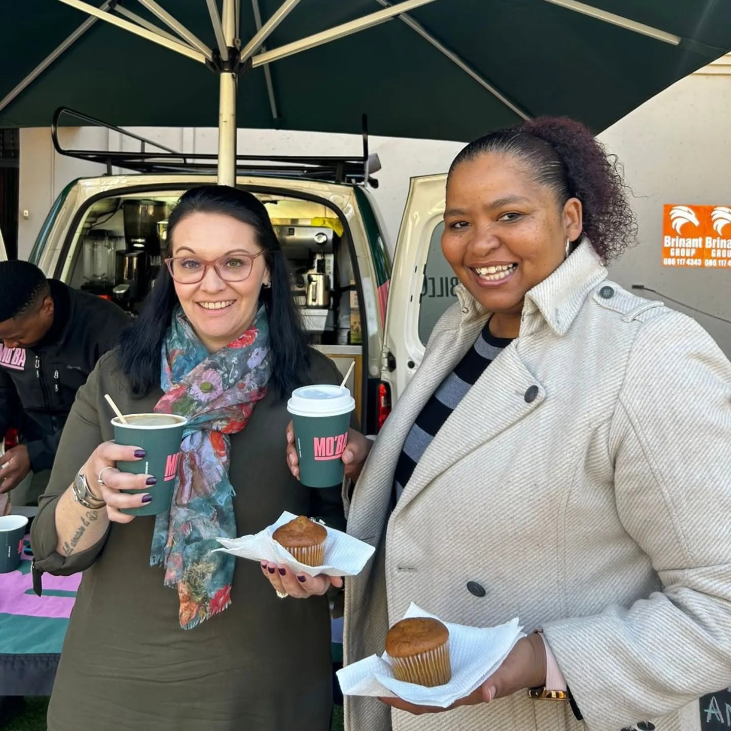 Two women smiling and holding MO'BA cups and muffins in front of the mobile bar.