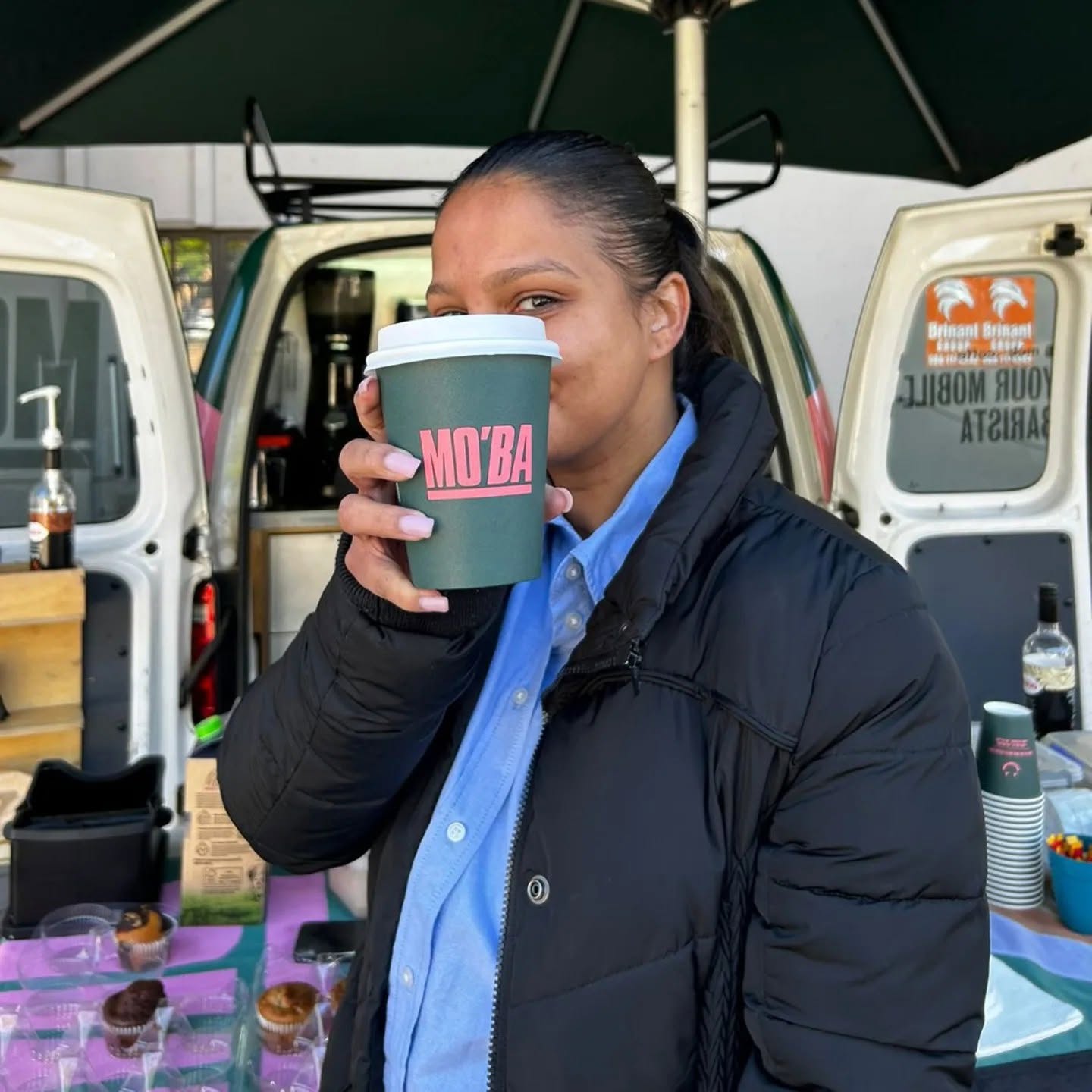 A woman taking a sip from a MO'BA cup, close up, with the van's mobile bar behind her.