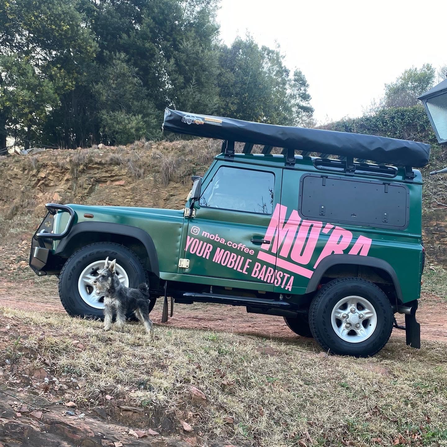 The MO'BA Land Rover Defender, wrapped in green with the pink MO'BA — Your Mobile Barista livery, parked on a dirt track in the South African countryside.